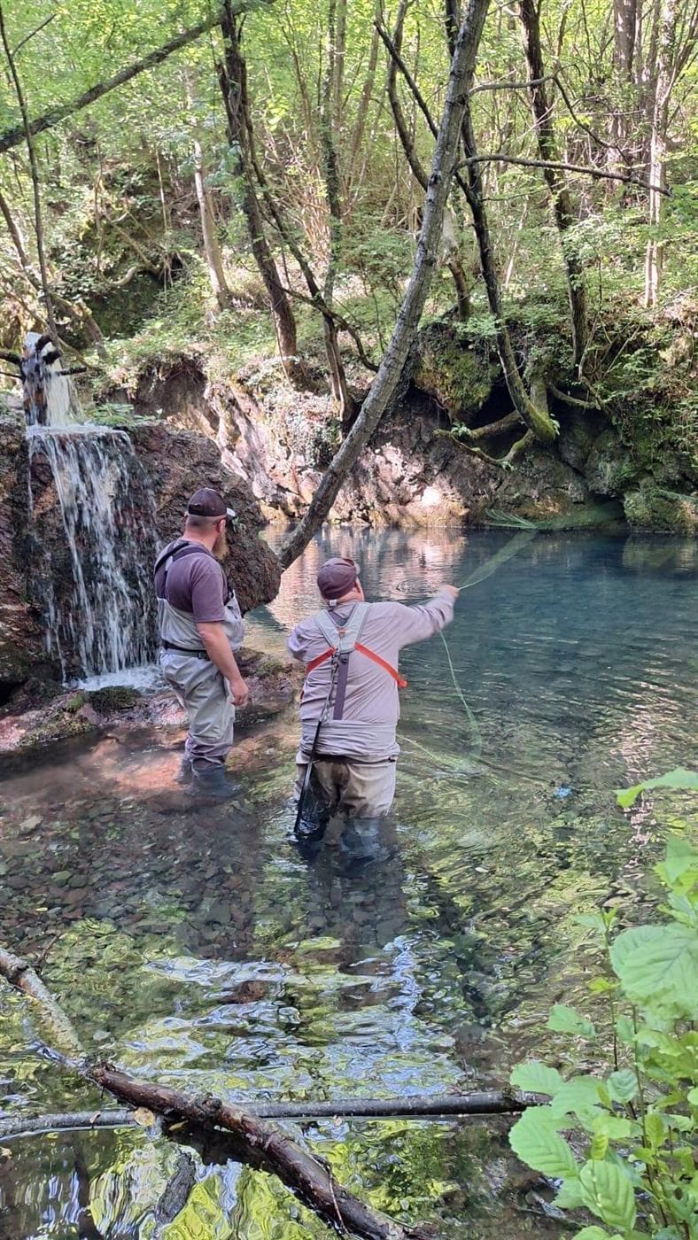 Two anglers wading a clear forest pool below a waterfall