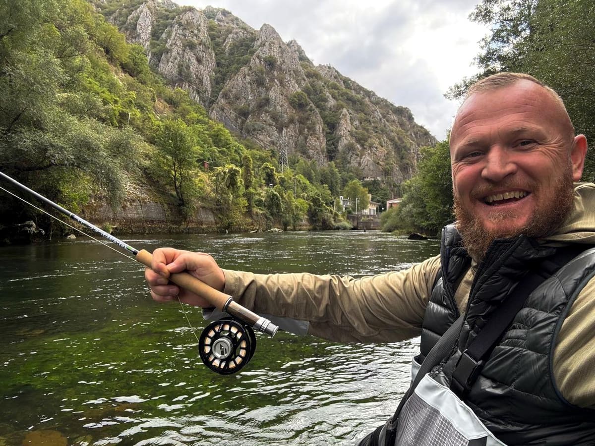 A smiling angler holding a fly rod on a river below rocky cliffs